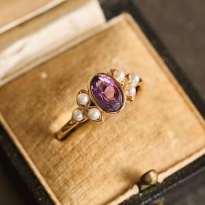 Könnte beinhalten: Ein goldener Ring mit einem ovalen, lila Edelstein, umgeben von kleinen, weißen Perlen. Der Ring wird in einer Vintage-Schmuckschatulle mit beigefarbenem Innenraum präsentiert. Das Design des Rings ist elegant.