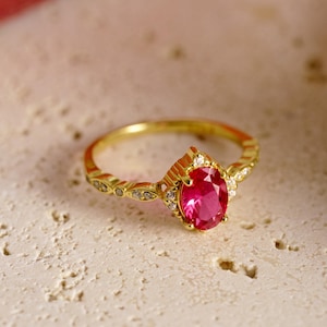 May include: A gold ring featuring an oval-cut, vibrant pink gemstone as the centerpiece. The ring's band is adorned with small, clear accent stones and delicate filigree detailing. The ring is set against a neutral, textured background.
