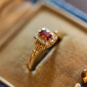 May include: A gold ring featuring a rectangular, deep red gemstone at its centre, encircled by smaller, clear stones. The ring has a decorative, floral-inspired design. The ring is presented in a blue and gold jewellery box.