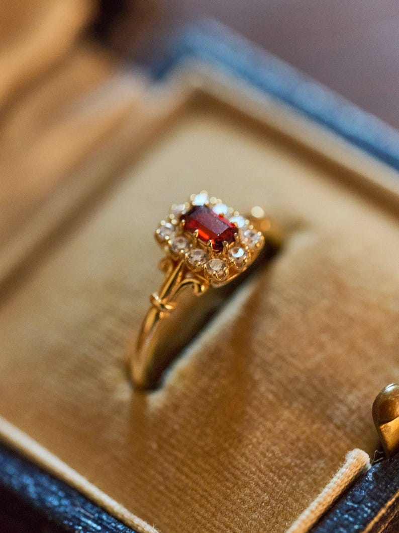 May include: A gold ring featuring a rectangular, deep red gemstone at its centre, encircled by smaller, clear stones. The ring has a decorative, floral-inspired design. The ring is presented in a blue and gold jewellery box.