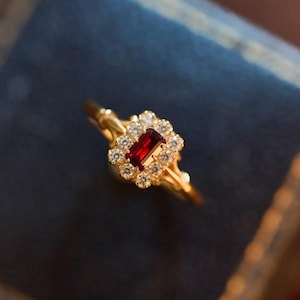 May include: A gold ring with a rectangular, crimson gemstone at its centre, encircled by a cluster of smaller, clear stones. The ring's band is slender and detailed. The setting has an ornate design, suggesting a vintage style. The ring is set against a dark blue backdrop.