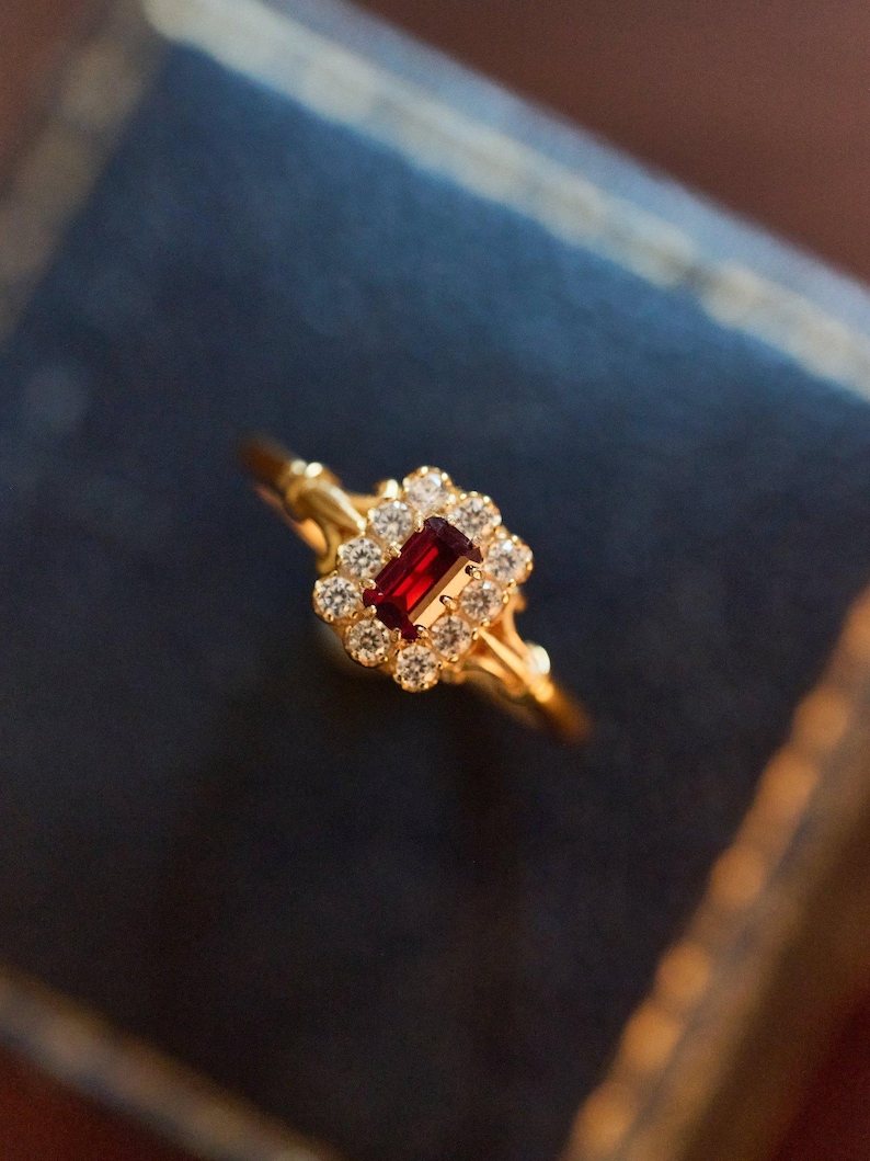 May include: A gold ring with a rectangular, crimson gemstone at its centre, encircled by a cluster of smaller, clear stones. The ring's band is slender and detailed. The setting has an ornate design, suggesting a vintage style. The ring is set against a dark blue backdrop.