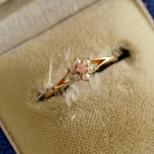 Op de afbeelding: Een delicate gouden ring met een bloemmotief, versierd met een centrale roze edelsteen en kleine parels. De ring wordt gepresenteerd in een blauwe sieradendoos met een lichtgele binnenkant, die de elegante details benadrukt.