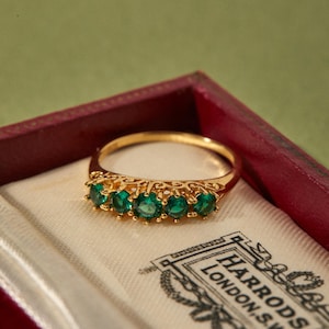 Puede incluir: Un anillo de oro con cinco piedras preciosas verdes talla esmeralda. El anillo está engastado en una banda de oro y se presenta en una caja roja con forro de satén blanco. La caja tiene el logotipo de Harrods London, S.W.