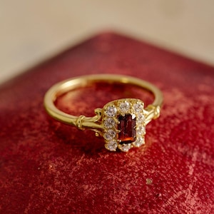 May include: A gold ring with a rectangular, deep red gemstone at its centre, encircled by smaller, clear stones. The ring has an intricate design and a slender band. The jewellery is set against a red backdrop, accentuating its features.