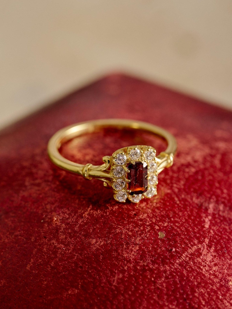 May include: A gold ring with a rectangular, deep red gemstone at its centre, encircled by smaller, clear stones. The ring has an intricate design and a slender band. The jewellery is set against a red backdrop, accentuating its features.