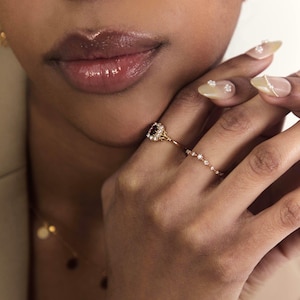 May include: A gold ring featuring a square, dark red gemstone encircled by small, clear stones. The ring is linked to a delicate chain of small, clear stones. The image also shows a gold necklace and a hand with decorated nails.