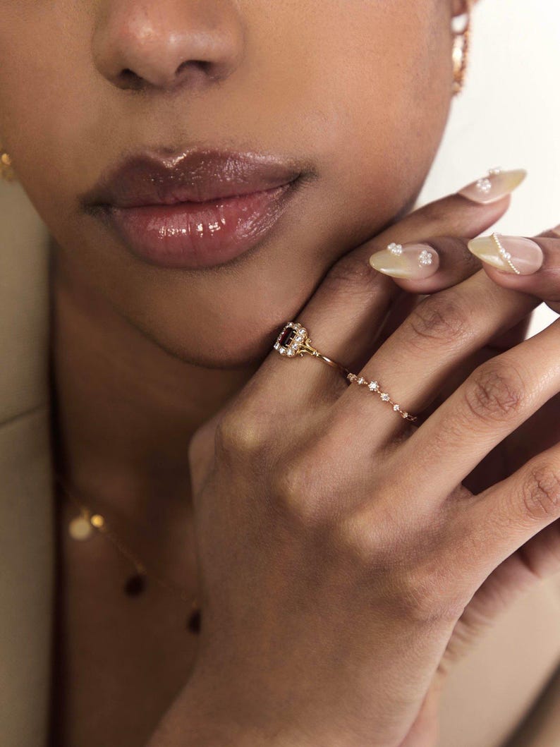 May include: A gold ring featuring a square, dark red gemstone encircled by small, clear stones. The ring is linked to a delicate chain of small, clear stones. The image also shows a gold necklace and a hand with decorated nails.