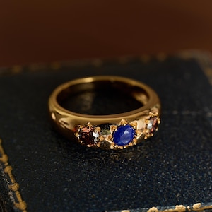 Op de afbeelding: Een gouden ring met drie edelstenen: een centrale blauwe steen geflankeerd door twee kleinere rode stenen. De ring heeft een gedetailleerde, stervormige zetting rond de blauwe edelsteen. De ring wordt gepresenteerd op een donkerblauwe ondergrond.