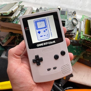 Puede incluir: Una consola de juegos portátil Game Boy Color blanca con una pantalla azul que muestra el texto "GAME BOY COLOR" y "Nintendo". La pantalla también muestra un juego pixelado azul y blanco. La consola tiene un pad direccional negro, botones negros y un altavoz.