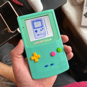 Puede incluir: Una consola de videojuegos portátil Game Boy Color de color turquesa con un pad direccional amarillo, botones rosa y verde, y una pantalla que muestra el texto "GAMECHANGER_MODS" y "GAME BOY COLOR".