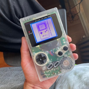 May include: A clear, transparent Game Boy handheld console with a screen displaying a purple graphic. The console has black buttons, a directional pad, and the text "DOT MATRIX WITH STEREO SOUND". The screen shows the text "GAMECHANGER.MODS".