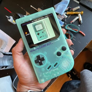 May include: A translucent teal Game Boy Color handheld video game console with a black and white screen displaying the text "SGAMECHANGER MODS". The console has a black and white button layout with the text "BATTERY", "Nintendo GAME BOY", "SELECT", and "START" printed on the console.