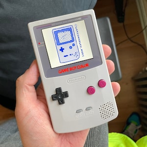 Puede incluir: Una consola de juegos portátil Game Boy Color blanca con una pantalla azul que muestra el texto "GAMECHANGER_MODS". La consola tiene un pad direccional negro, botones negros y botones A y B rosas.