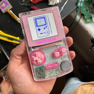 Puede incluir: Una consola de videojuegos portátil Game Boy Color rosa con una carcasa de plástico transparente. La consola tiene una pantalla azul y blanca con el texto "GAMECHANGER_MODS" y "GAME BOY COLOR" en ella. La consola tiene un pad direccional rosa y botones rosas.