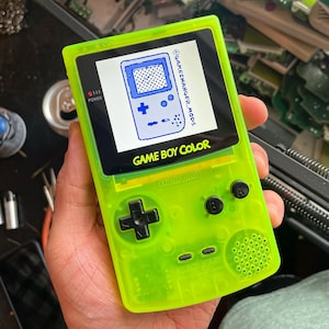 Puede incluir: Una consola de juegos portátil Game Boy Color de color verde neón. La pantalla muestra un dibujo lineal azul de una Game Boy. La consola tiene botones negros y el texto "GAME BOY COLOR" es visible. El logotipo de Nintendo también es visible.