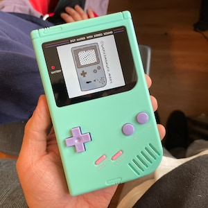 May include: A mint green handheld gaming console with a screen displaying a retro game graphic. The console features a purple directional pad and buttons, with the text "DOT MATRIX WITH STEREO SOUND" and "GAMECHANGER.MODS" visible on the screen.