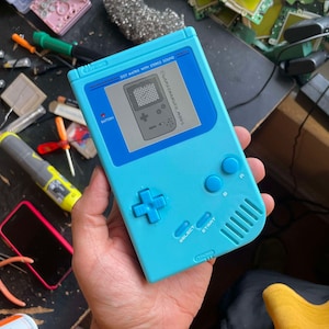May include: A blue Game & Watch: Super Mario Bros. handheld game console with a black and white screen. The console has a red button labeled "BATTERY" and a white button labeled "POWER". The console has a white directional pad, four white buttons, and a white "SELECT" button. The console has a white "START" button. The console has a white label that reads "GAME & WATCH: SUPER MARIO BROS.". The console has a white label that reads "DOT MATRIX WITH STEREO SOUND".