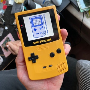 Puede incluir: Una consola de videojuegos portátil Game Boy Color amarilla con una pantalla azul que muestra un juego. La pantalla tiene el texto "GAMECHANGER-MODS" escrito en ella. La consola tiene botones negros y un altavoz.