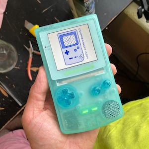 Può includere: Un Game Boy Color blu traslucido con uno schermo blu e bianco. Lo schermo mostra il testo "GAMECHANGER MODS" e "GAME BOY COLOR". Il dispositivo ha un pulsante a croce blu, due pulsanti rotondi blu e una luce verde.