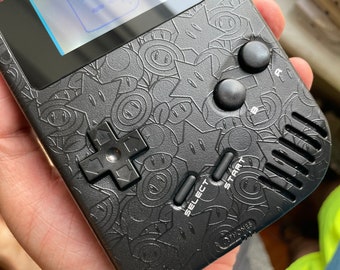 Può includere: Console di gioco portatile nera con un motivo a stelle e facce sorridenti. Dispone di un D-pad, pulsanti select, start e A/B. Lo schermo è visibile nella parte superiore.
