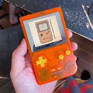 Puede incluir: Una Game Boy Pocket transparente naranja con botones amarillos y una pegatina en la pantalla que dice "Gamechanger Mods" y "Limited Edition (Model 1)".