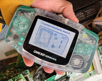 Puede incluir: Una consola de juegos portátil Game Boy Advance transparente, clara y translúcida, con un diseño verde y blanco. La pantalla muestra el texto "Game Boy Advance" y "GameChanger_Mods".