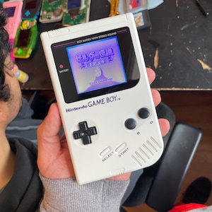 May include: A white Nintendo Game Boy handheld gaming console with a black and white screen displaying the game "The Legend of Zelda: Link's Awakening DX". The console has a red dot on the battery indicator, and the text "DOT MATRIX WITH STEREO SOUND" is printed on the top of the console.