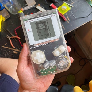 May include: A clear, transparent Game Boy handheld gaming console with a black and white screen. The screen displays the text "GAME & WATCH" and "GAME BOY". The console has a white cross-shaped button and two white round buttons.