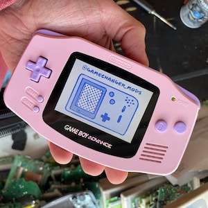 以下が含まれることがあります： 紫色のボタンセットが付いたピンクのゲームボーイアドバンス携帯ゲーム機。画面には、「@GAMECHANGER_MODS」というテキストが表示された青と白のピクセル化された画像が表示されています。