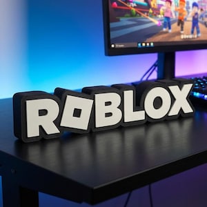May include: A black desk with a computer monitor displaying a Roblox game. The word "ROBLOX" is displayed in white block letters with a black outline. A keyboard and mouse are also on the desk.