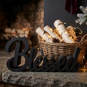 Puede incluir: Un letrero de madera negro con la palabra "Blessed" en cursiva se encuentra sobre una superficie de piedra. Una cesta de mimbre llena de troncos de abedul, pi&ntilde;as y luces de hadas se encuentra en el fondo, cerca de una chimenea.