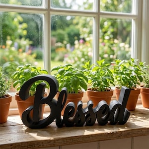 Puede incluir: Un letrero de madera negro con la palabra "Blessed" est&aacute; colocado en un alf&eacute;izar de madera. Detr&aacute;s, varias plantas en macetas con hojas verdes est&aacute;n dispuestas, con una ventana que ofrece vistas a un jard&iacute;n.