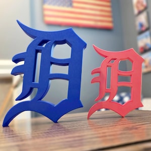 May include: Two stylized letter "D" sculptures, one blue and one red, stand on a wooden surface. The "D"s have a classic, bold font with a distinctive serif design. The background includes an American flag.