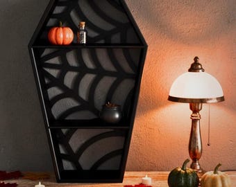 May include: A black coffin-shaped shelf with a spiderweb design, holding an orange pumpkin, a small glass bottle, and a black cauldron. A vintage lamp and pumpkins sit on a wooden surface with autumn leaves. Halloween decor.