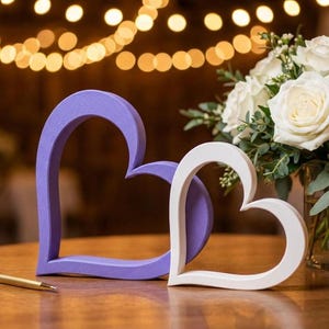 May include: Two heart-shaped decorations, one lavender and one white, sit on a wooden surface. A gold pen rests nearby. A bouquet of white roses in a glass vase is in the background, with soft bokeh lights.