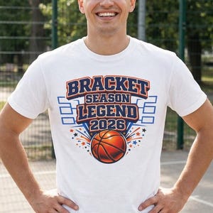 May include: White t-shirt with the text "BRACKET SEASON LEGEND 2026" in blue and orange. A basketball graphic is centered below the text. The shirt is worn by a person standing outdoors.