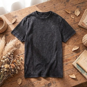 Puede incluir: Una camiseta gris oscuro, estilo lavado al ácido, con cuello redondo y mangas cortas. La camiseta está colocada sobre una superficie de madera rústica, rodeada de flores secas, libros y cestas decorativas. La estética general es informal y vintage.