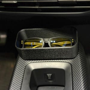 May include: Black rectangular sunglasses with yellow lenses rest in a black tray inside a car. The tray is placed in the center console, near the air vents. The console has a carbon fiber pattern.