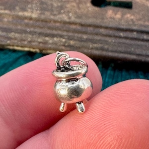 May include: A silver charm in the shape of a cauldron with a handle. The cauldron has three legs and a textured surface.