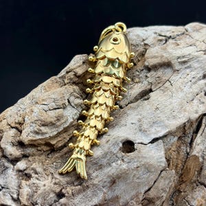 May include: A gold-toned metal pendant shaped like a fish with articulated segments. The fish features a smooth head with a small circular eye, a body covered in overlapping scale-like plates, and a flared tail fin. A small loop is at the top.