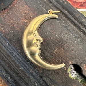 May include: A gold-toned crescent moon charm with a face sculpted into the surface. The charm is on a dark wooden surface.