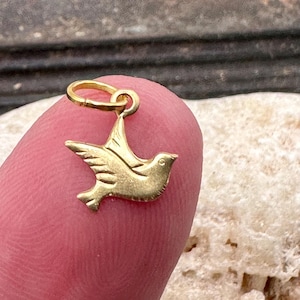 May include: Gold-toned metal charm in the shape of a dove. The dove is in flight with its wings spread. The charm has a loop at the top for attaching to a necklace or bracelet.