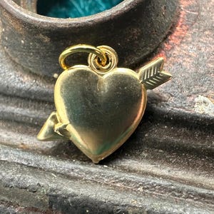 May include: A gold heart charm with an arrow through it. The charm is on a rustic brown surface.