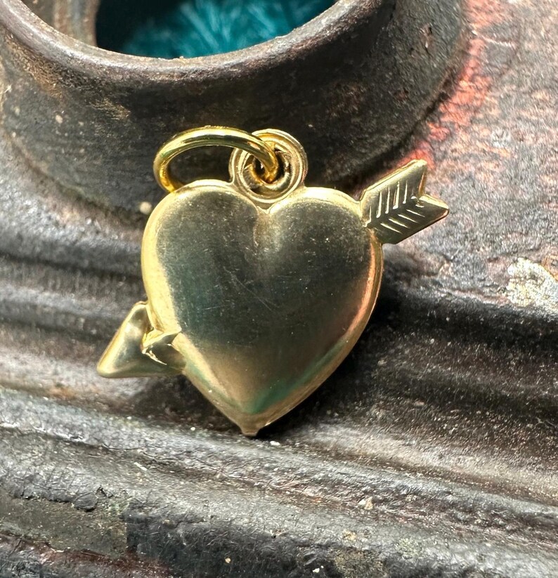 May include: A gold heart charm with an arrow through it. The charm is on a rustic brown surface.