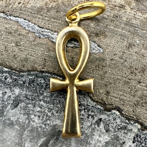 May include: A gold-colored Ankh pendant with a loop at the top for a chain. The Ankh symbol, a cross with a teardrop-shaped loop, is a symbol of life. The pendant is displayed against a textured, gray background.