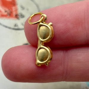 Puede incluir: Un pequeño dije dorado con forma de gafas. El dije tiene una anilla redonda en la parte superior para sujetarlo a una cadena o pulsera. Las gafas tienen lentes ovaladas y un diseño de montura clásico.