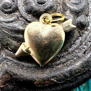 May include: Gold heart charm with an arrow through it. The charm has a loop for attaching to a necklace or bracelet.