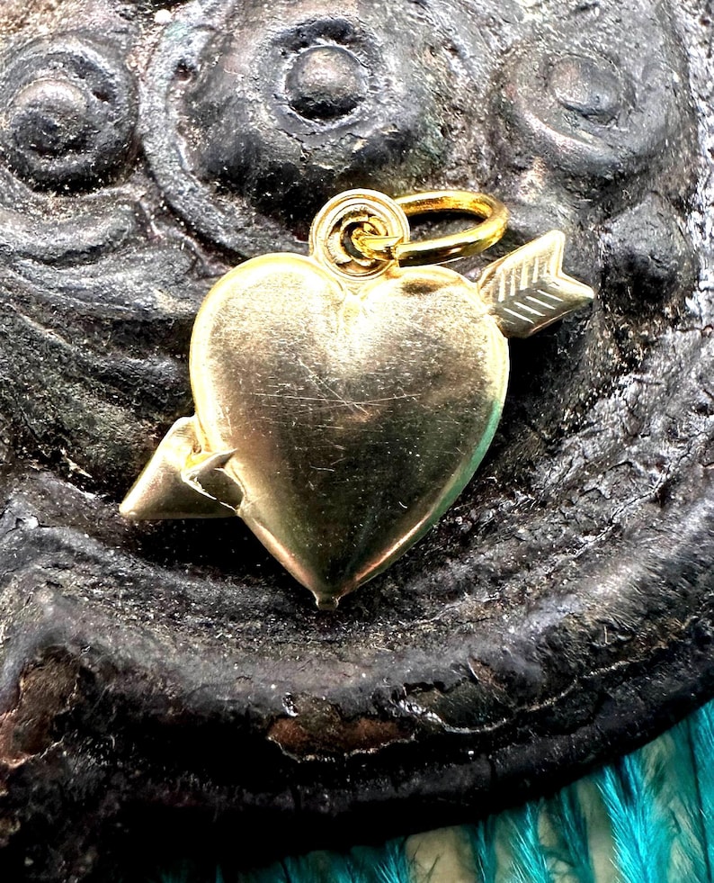 May include: Gold heart charm with an arrow through it. The charm has a loop for attaching to a necklace or bracelet.