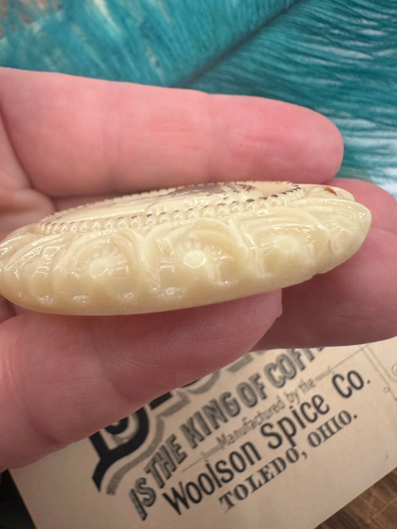 May include: A white ceramic button with an intricate design. The button has a raised, floral pattern around the edge. The text "IS THE KING OF COFFEE" is printed on a label below the button. The label also includes the text "Manufactured by the Woolson Spice Co. TOLEDO, OHIO."